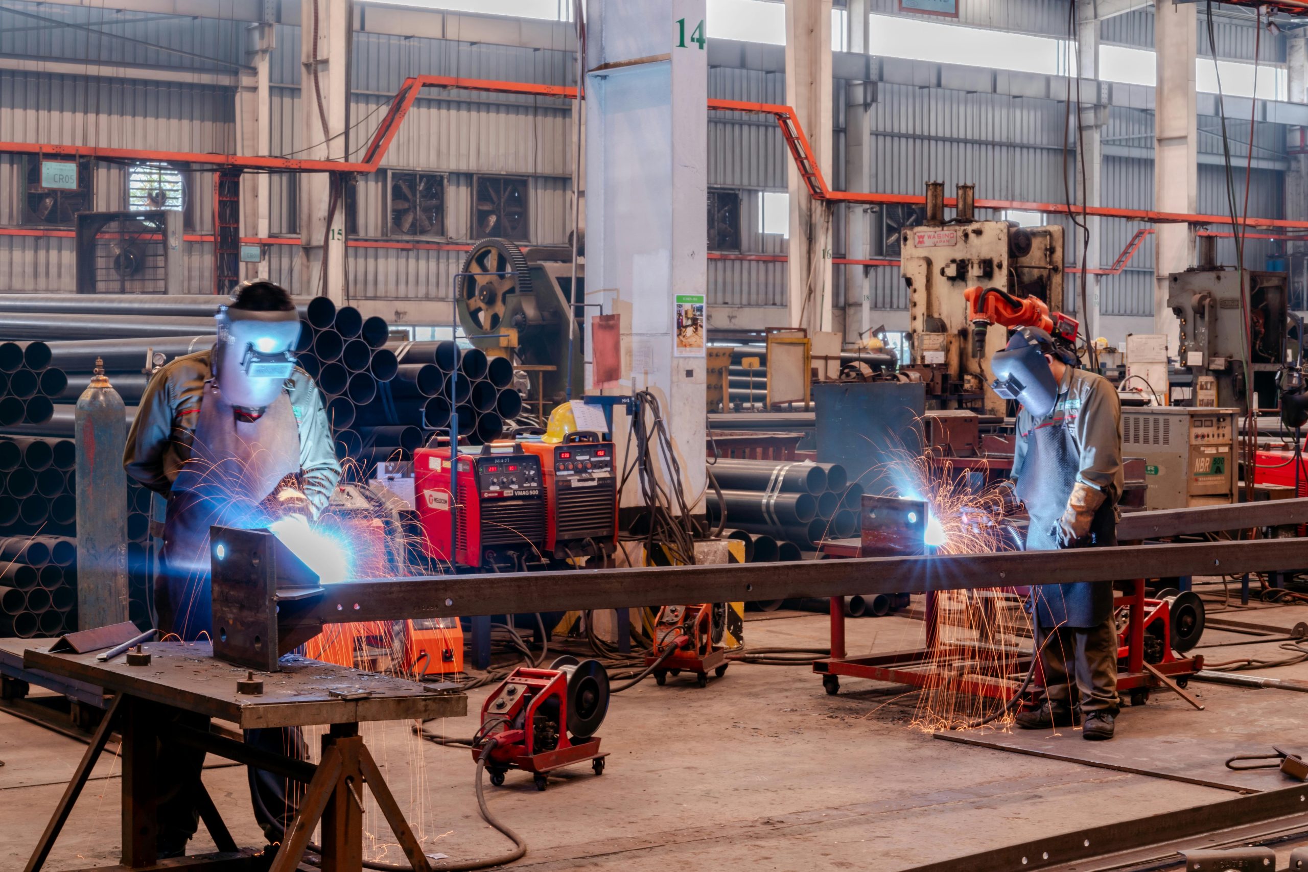 Two welders wearing protective gear working in a busy industrial factory workshop.